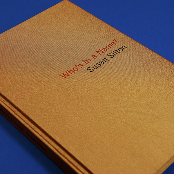 This artwork by the artist Susan Silton depicts the front cover of a tan bound book with red text that reads Who's in a Name? and black text that says Susan Silton. The book lays flat and angled against a deep blue background surface.