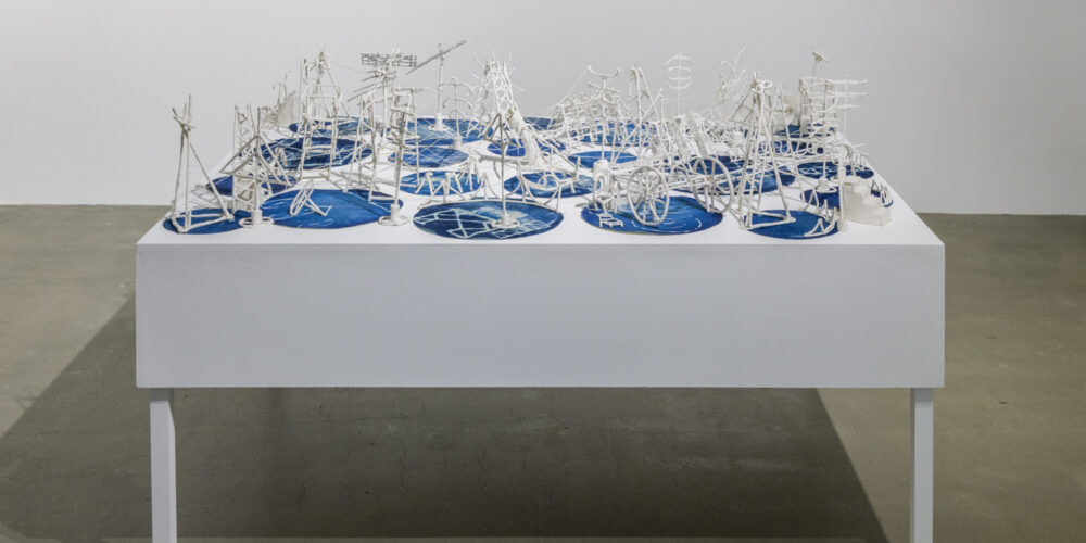 This artwork installation by the artist Fran Siegel features various white ceramic structures on circular, cyanotype prints. These are all placed on top of a white table.