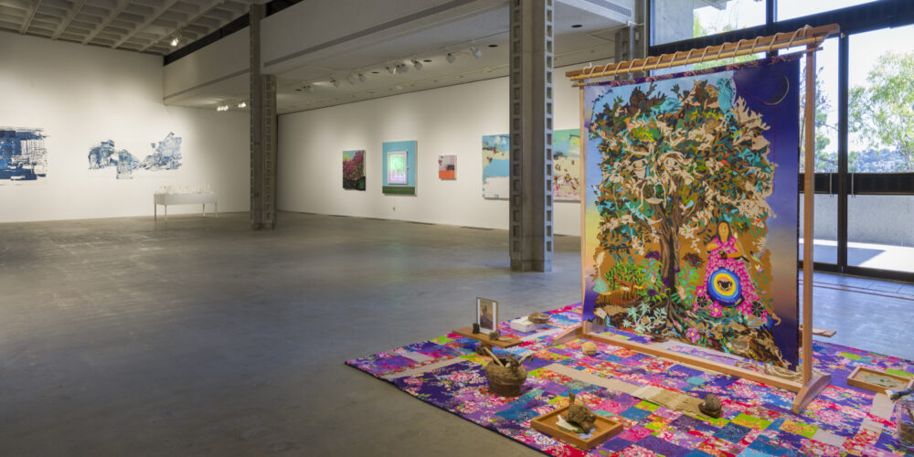 In this art installation, there is one large artwork by the artist iris yirei hu that features a floral textile with pink, purple, blue and yellow colors on the floor. There are also various objects, such as a pile of dirt, a woven basket and a picture frame placed on the textile. Standing in the middle of the textile is a large freestanding tapestry that depicts a woman wearing a bright pink robe and standing next to a tree. To the left of this artwork installation are artworks by the artist Patrick Martinez, including a neon image of a palm tree, a small purple and orange painting, and another painting of bougainvillea. To the left of these artworks is a different artwork installation by the artist Fran Siegel. These artworks include white and blue abstract paperworks hanging on the wall, and a white table with white ceramic, abstract forms sitting on top.