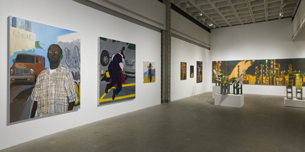 The artwork installation by the artist Henry Taylor features three paintings of various sizes depicting different figures. One painting depicts a male, person of color wearing a light colored checkered shirt with a brown van parked behind him over his left shoulder. To the right of this painting are two figures (a white police officer and a person of color) in a struggle on the floor. To the right of this artwork is a small painting of a male, person of color sitting in a brown chair. To the right of this artwork installation is another installation by a different artist named Mario Ybarra Jr. This artwork installation includes a large, horizontal painting of the cityscape of Wilmington in black and orange colors, photographs of construction workers, and medium-sized wooden replicas of cranes placed on top of white pedestals.