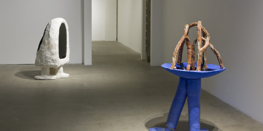 The two artwork sculptures by the artist Anna Sew Hoy depict amorphous figures. One of the figures is blue with three legs with a bird bath on top, and the other figure has two legs and a black mirror front.