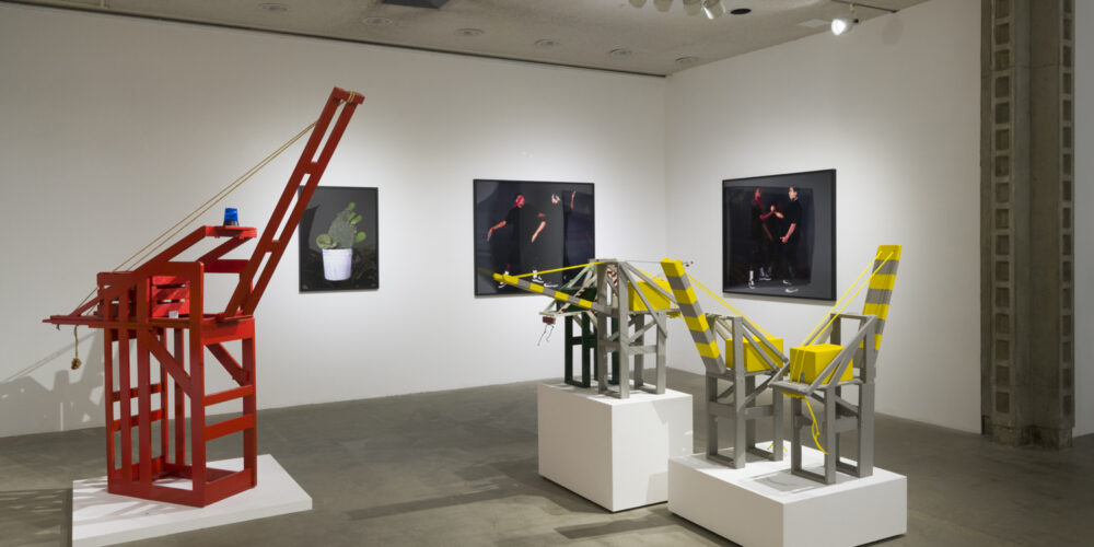 In this artwork installation by the artist Mario Ybarra Jr., there is one large red, wooden sculpture depicting a crane, with two smaller wooden sculptures depicting cranes (painted grey and yellow) to the red crane's right. There are also three photographs hanging on the wall behind these sculptures; one depicts a cactus, another photograph depicts two figures of color engaged in a knife fight, and the other photograph depicts the same two figures clasping hands in a friendly manner.