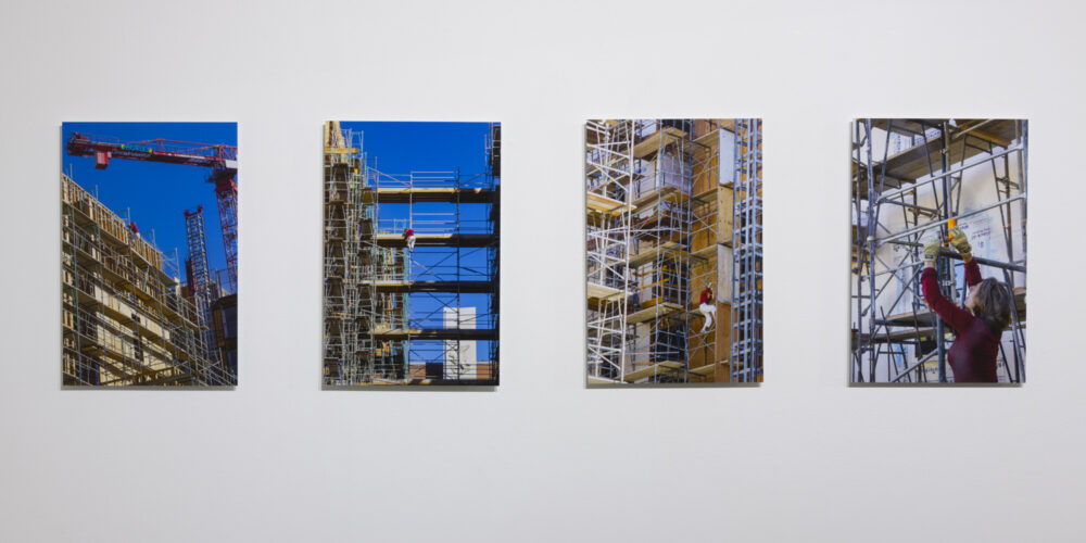 In this artwork installation by the artist Nancy Popp, there are four photographs of different construction sites. In each of the photographs, the artist is depicted in the midst of tying orange rope in and around the construction sites.