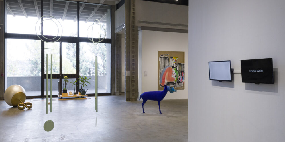 There are two light green sculptural chime works hanging from the ceiling by the artists Rebecca Bruno and Mak Kern. Behind this work to the left is a large sculpture depicting a pearl stud placed on the floor by the artist Alex Nazari. To the right of this artwork is an installation of plumeria plants on a wooden dolly by the artist David Horvitz. To the right of this work is a large abstract painting with various shades of pink, black, blue and green on brown canvas by the artist Lindsay August-Salazar. In front of this artwork is a sculpture depicting a cobalt-blue deer with a plastic chips bag over its head by the artists Galería Perdida. And in front of this work is a video artwork on two TV monitors by the artists pecking nets. One TV monitor has a blank white screen and the other TV monitor has a black screen with the words Spatial White.