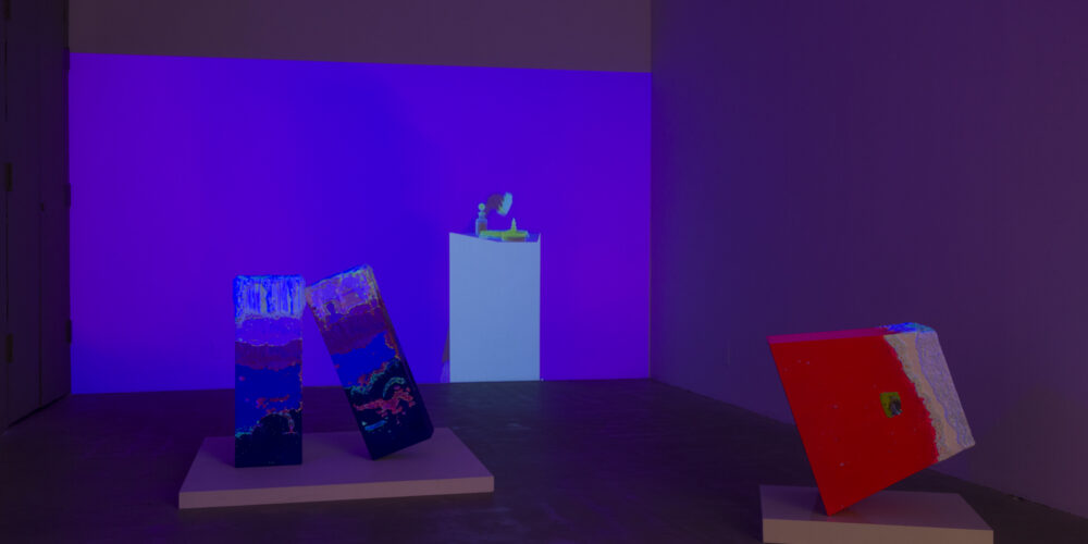 In this artwork installation by the artist Lena Daly, a flourescent, neon colored video projection is displayed on a wall. This video projection still features an image of a small sculptural work on a pedestal.