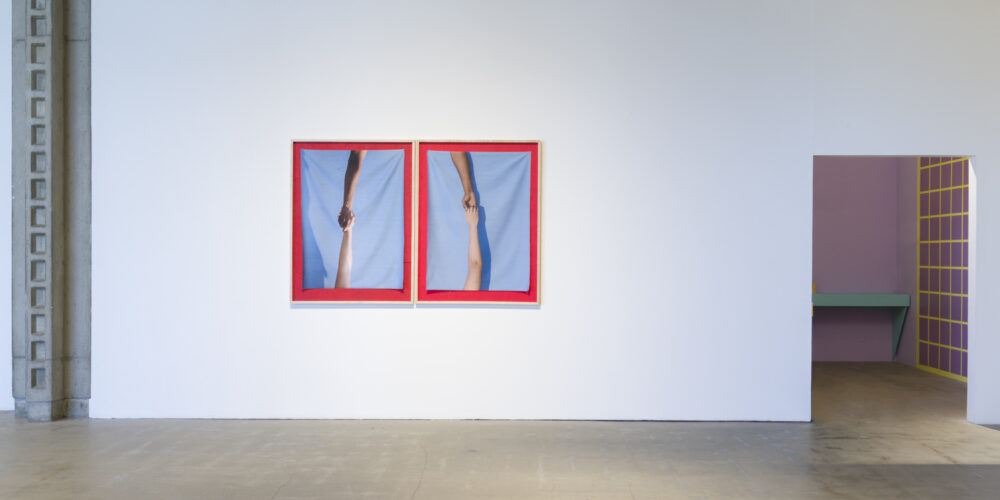 In this artwork installation by the artist David Alekhuogie, two artworks are hanging on the wall and in a diptych format. One photograph depicts an arm from a white individual and an arm from a person of color clasping hands. These arms are situated vertically, so the person of color arm is on top. There is a light blue background for these arms and a red outline surrounding the image. The other artwork resembles this artwork. There is also another artwork placed on the floor in front of this diptych, which depicts a blue bandana with red paint splattered on it.