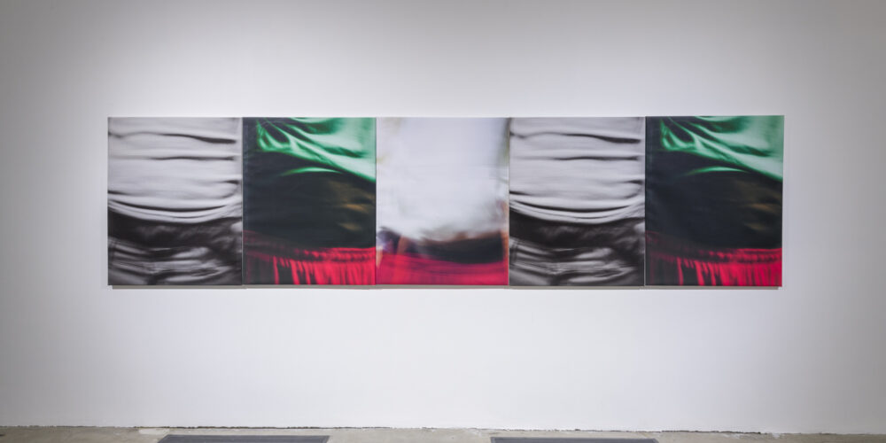 This is an artwork installation by the artist David Alekhuogie featuring five photographs printed on canvas. Each of these photographs depict slightly different angles of figures wearing saggy pants and in different colors. The far left photograph depicts a figure wearing a crinkled white top and grey crinkled pants viewed mid torso to the top of the pants, while the photograph to the right of this work depicts another figure wearing a green top and red elastic-waisted pants viewed mid torson to the top of the pants; the green top is pulled up higher than the other photograph. These two photographs are also shown in reverse order (photograph of the figure wearing a green top and red elastic-waisted pants on the far right) on the right side. In the middle of these two suites of photographs is another photograph of a blurry figure wearing red elastic-waisted pants. The top portion is white with no fabric wrinkles or creases visible. These five artworks are placed side-by-side in a row.