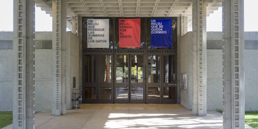 There are three artworks hanging from the ceiling by David Horvitz facing the doors outside of the art gallery. The left sign is white with black lettering that says "un reloj que persigue las sombras de los gatos". The middle sign is red with black lettering that says "un reloj reido por el viento". The right sign is blue with white lettering that says " un reloj que se queda dormido".