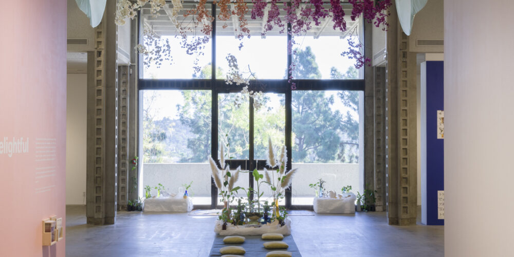 The artwork installation by the artist Lani Trock includes a long blue rug with eight green colored floor pillows separated in two lines. There are three pillows on the left side, four pillows on the right side, and one pillow in the middle. There are various green and white colored plants in glass vases placed on the back of the rug. There are more green plants in glass vases placed on a short, white rectangular pedestal to the left back of the rug and to the right back of the rug. Above the rug and hanging from the ceiling are rows of flowers. The left side has white flowers, the middle has orange flowers, and the right has purple flowers. There are also white and light blue colored fabric hanging with the flowers as part of the artwork installation.