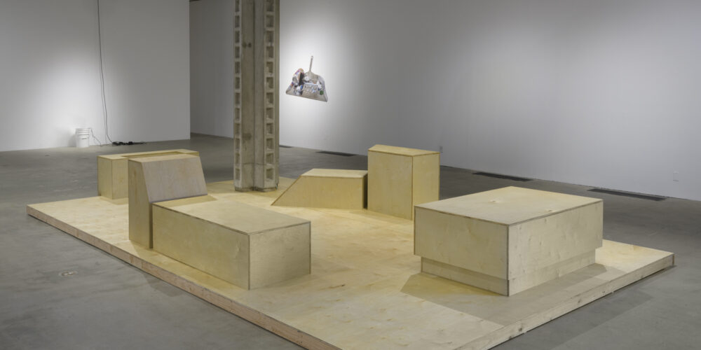 The artwork installation by the artist Lauren Davis Fisher includes five plywood box sculptures shaped with various geometric forms and dimensions. They are placed as seating arrangements on top of a large rectangle plywood platform. To the left this artwork installation is another artwork by the artist Dylan Mira of a small white bucket placed on the floor. There is mist coming out of the bucket and power cords trailing out of it and up along the wall. To the right of this artwork, there is another artwork hanging on the wall by the artist Asha Schechter. This artwork depicts a dustpan with various objects.