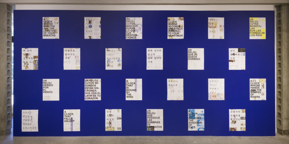 The art installation by the artist David Horvitz is comprised of a large, cobalt blue painted wall with twenty-four paper posters separated into four rows of six posters per row. The posters are hanging vertically and have various black lettering in different languages such as English, Spanish, Japanese and Korean.