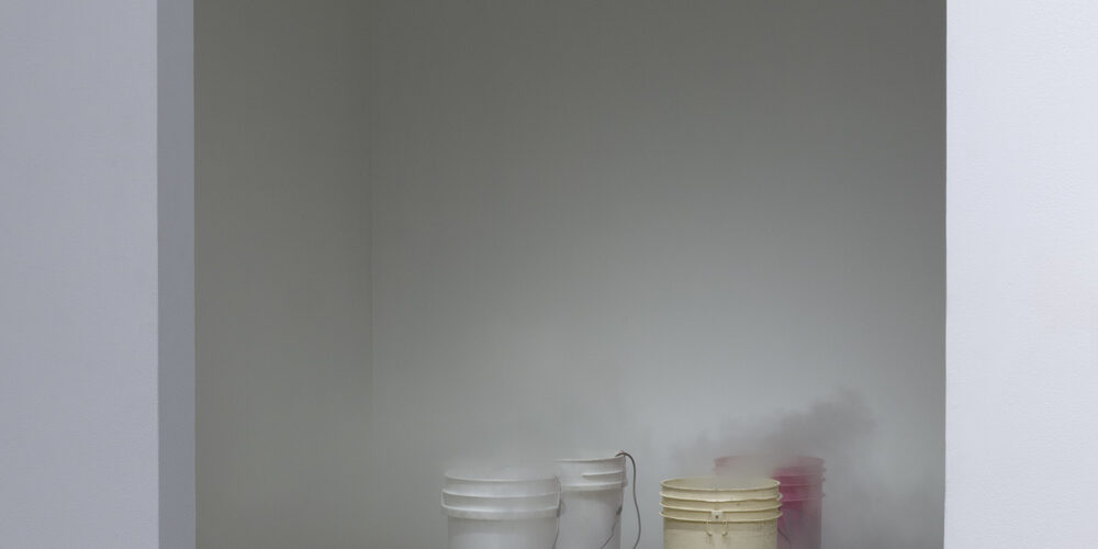 The art installation by the artist Dylan Mira is comprised of four paint buckets arranged in a square shape on the floor. The front row has one white bucket on the left and one yellow bucket on the right. The back row has one white bucket o the left and one pink bucket on the right. There is mist coming from the tops of the buckets and spilling out on to the floor.