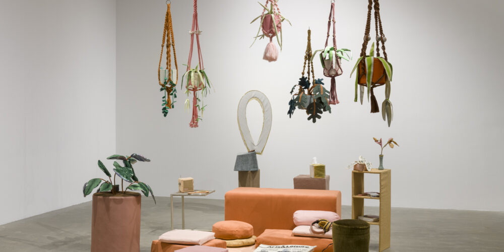 various sized square cushions in shades of pink, orange and red. On top of one of the pink floor cushions is a folded, fabric newspaper, and on another pink pillow is a pair of fabric headphones. Behind the rug is an orange colored bench, and behind the bench is a fabric sculpture in the shape of a circular ring and another fabric pedestal with an object in the shape of a Kleenex box. To the left side of the circular pink mat is a large fabric bird of paradise plant in a purple colored fabric planter. To the right of the circular pink mat is a brown fabric stool and a fabric side table with two shelves that have various fabric objects. There are six fabric plants hanging above the circular pink mat in various shades of pink, orange and brown hanging planters.