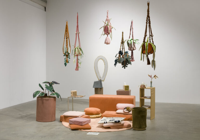 various sized square cushions in shades of pink, orange and red. On top of one of the pink floor cushions is a folded, fabric newspaper, and on another pink pillow is a pair of fabric headphones. Behind the rug is an orange colored bench, and behind the bench is a fabric sculpture in the shape of a circular ring and another fabric pedestal with an object in the shape of a Kleenex box. To the left side of the circular pink mat is a large fabric bird of paradise plant in a purple colored fabric planter. To the right of the circular pink mat is a brown fabric stool and a fabric side table with two shelves that have various fabric objects. There are six fabric plants hanging above the circular pink mat in various shades of pink, orange and brown hanging planters.