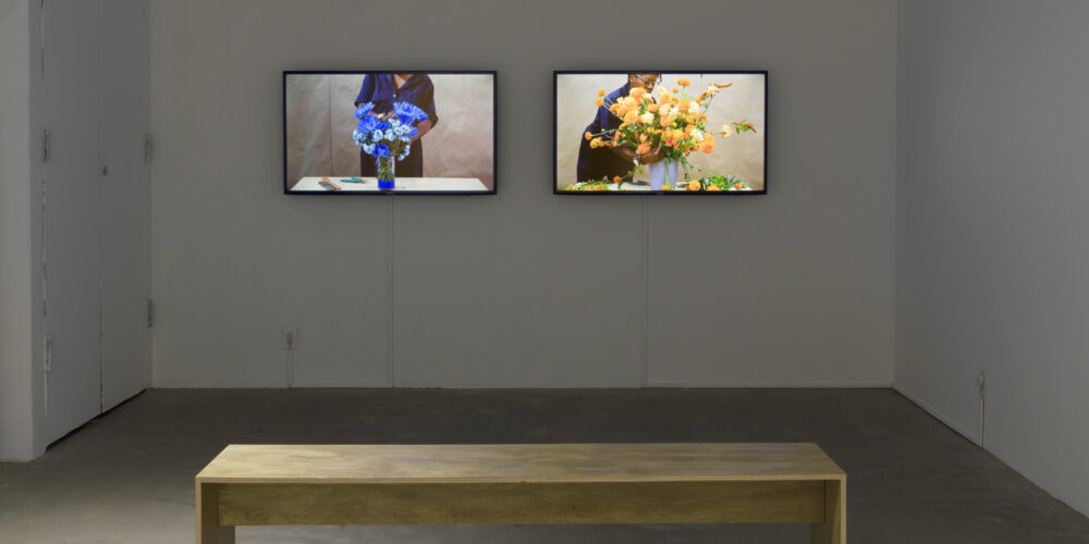 The video art installation by the artist Cauleen Smith is featured on two different television screens. On the left screen is a woman making a blue colored flower arrangement. On the right screen is the same woman making an orange colored floral arrangement. Both videos are by the artist Cauleen Smith.