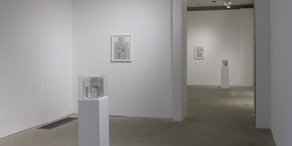 In this art installation by the artist Milano Chow, there is one artwork on a white pedestal with a plexiglass cover. Under the cover is a small graphite sculptural work featuring an architectural space and an upright partition with a figure peeking out from behind it. Behind this artwork is another graphite drawing of a different architectural space hanging on a wall. There are two other similar artworks placed in the next room behind these works.