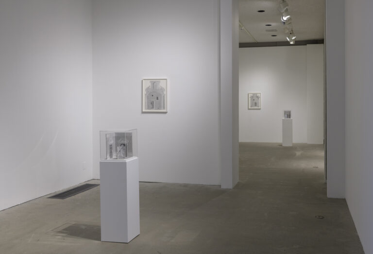 In this art installation by the artist Milano Chow, there is one artwork on a white pedestal with a plexiglass cover. Under the cover is a small graphite sculptural work featuring an architectural space and an upright partition with a figure peeking out from behind it. Behind this artwork is another graphite drawing of a different architectural space hanging on a wall. There are two other similar artworks placed in the next room behind these works.