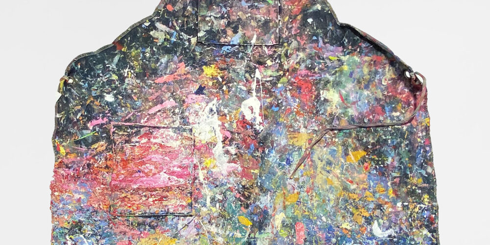 This is a color photograph of an apron owned by Vanessa McConnell. The apron is made of a dark-colored fabric, and it is covered in brightly colored paint splatters and strokes. The paint colors include pink, red, yellow, green, orange, blue, and white.