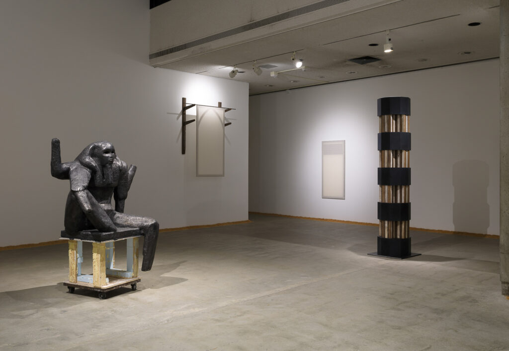 This is a color photograph of five artworks by Lukas Geronimas. On the left in the foreground is the sculpture Puzzlemaster, which is a sculpture of a dark grey seated figure on a wood pedestal placed on top of a dolly. To the right of this work is the sculpture Custom Hung Valance, which is a sculpture constructed of wooden racks and stretched muslin resembling a Gothic-style window. To the right of this work on a perpendicular wall in the background is the artwork Custom Vent, which is hung sculpture made of muslin and plywood resembling a vent on a wall. On the far right in the middle ground is the sculpture Barnsdall Column, which is constructed in nine segments with four multi-paneled and colored wood components in the shape of a cross and five ten-sided prisms made of a custom resurfaced plexiglass in a dark grey-black color. The cross pieces are modeled after the LAMAG's unique column design. Along the perimeter of the floor and walls of this installation is another artwork titled Puzzledust, which is sawdust that was produced, collected and stored by the artist over the past two years.