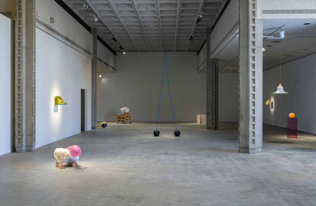 This is a color photograph of an installation view of artworks by Nevine Mahmoud. Pictured left to right are: a sculpture titled HOT TOY STRADDLE, which is a large white boulder with a pink object placed on top and sitting on the floor; a sculpture titled Bust (lagoon) installed on the adjacent wall, which resembles a bare female chest made from hand blown glass and resin in a yellow-green color; a sculpture titled Beach Ball IV in the background, which is a spherical ceramic work, cracked in a few places, painted with pink, yellow, red, and blue glaze made to appear like an inflated beach ball; a sculpture titled Three Components, which is a large white boulder with a light pink object placed on top and sits on top of a wooden crate; a sculpture titled Aileen & Elvis, which resembles a two-stemmed black cherry with blue stem; a sculpture titled Castle component #1, which resembles two, light pink colored puzzle pieces criss-crossed; a sculpture installed on the adjacent wall titled Babette, which resembles a bare female chest in a peach-tan skin color made from hand blown glass, resin, and aluminum; a sculpture titled Sherry which depicts a bright orange-red colored cherry shaped sculpture placed on top of a fuschia colored plexiglass pedestal; and a hung sculpture titled Breast shade, which resembles a bare female breast made from alabaster and pigmented resin.