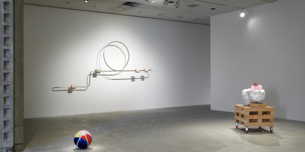 This is a color photograph of three sculptures by Nevine Mahmoud. Pictured left to right are: a sculpture titled Beach Ball IV, which is a multi-colored spherical ceramic work placed on the floor and resembles an inflatable beach ball; a wall-mounted sculpture titled Abacus arm 1, which features a linear steel bar with a circle and four stone components. Each of these components are wrapped around the steel bar and are different stones, such as onyx, Portuguese marble and Aphrodite marble; and a sculpture titled Three Components, which includes a large white boulder with a small, pink, bulbous glass object that has a red coil placed on top of it. These particular elements sit on top of a large stack of wood pieces on wheels.