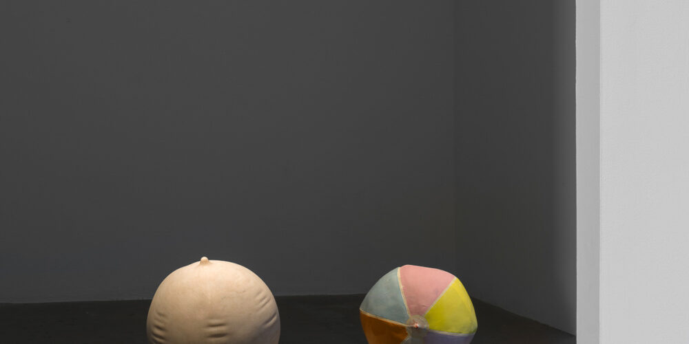 This is a color photograph of two sculptures by Nevine Mahmoud. The sculpture on the left is Ball Stripped Bare, which is spherical, made of Portuguese pink marble, and resembles an inflated beach ball. To the right of this work is Deflating Beach Ball II, which is slightly smaller in comparison to the other work and is made of ceramic with a multicolored in colors of pink, light blue, yellow and orange glaze mimicking the look of a slightly deflated beach ball. Both sculptures are placed directly on the floor and side-by-side.