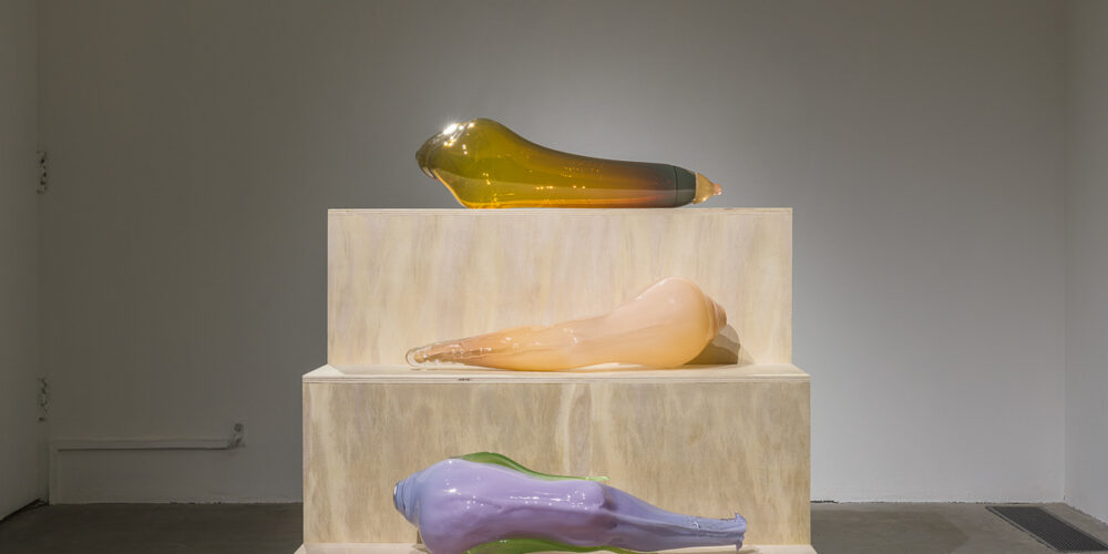 This is a color photograph of three large blown glass sculptures of roughly equal size by Nevine Mahmoud resting on a tiered, wooden three step pedestal. At the top is a sculpture entitled yellow Toy, an amber colored blown glass piece resembling a human leg. On the center tier is a sculpture entitled Leg (Lila), a flesh toned blown glass piece resembling a human leg. Beneath this on the last tier is another leg shaped work called Leg (lilac), made of lilac and green colored blown glass.