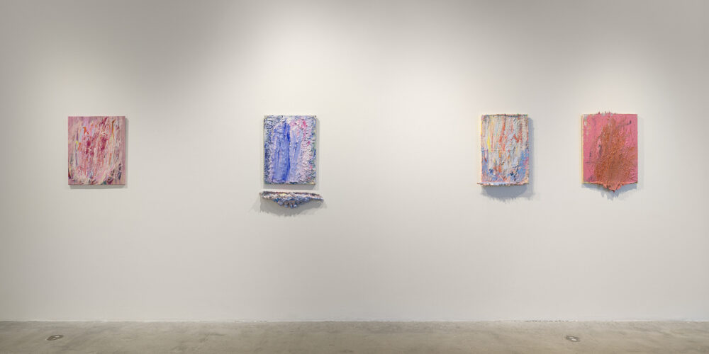 This is a color photograph of a white gallery wall with four hung artworks by Vanessa McConnell. Each artwork is an abstract painting of roughly equal size. On the far left is a painting utilizing mostly white, pink, and red paint, applied thickly to the canvas in mostly vertical strokes. In the left center is a painting consisting of mostly blue shades of paint with some pink and red as well, applied thickly to the canvas. A separate irregularly shaped piece in a similar color scheme is hung directly underneath. In the right center is a painting consisting of various colors, including yellow, blue, and red in mostly vertical strokes. On the far right is a painting featuring thick, textured layers of pink and orange paint, which extends and stretches outward from the bottom of the canvas.