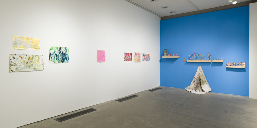 This is a color photograph of two walls in an exhibition of Vanessa McConnell's artworks at the Los Angeles Municipal Art Gallery. On the right white wall are seven brightly colored abstract paintings on paper by Vanessa McConnell. On the far right is a grouping of three, two featuring the use of yellow and green paint mounted one on top of the other on the left, and on the right a third painting featuring mostly blue and green paint. On the center of the left wall is a painting featuring primarily pink paint. On the right side of this wall is another grouping of three paintings, featuring mostly shades of red, yellow, and blue paint. On the right blue wall are four floating shelves containing a variety of paint-covered objects, including paintbrushes and various cups and dishes. In the center of the wall underneath one of the shelves is a paint splattered fabric drop cloth, draped on a single hook flowing down the wall and resting on the floor.