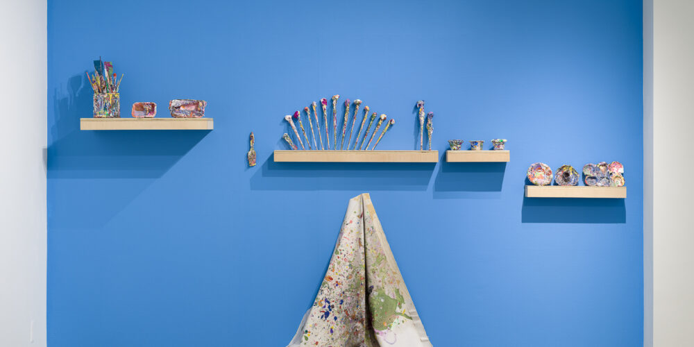 This is a color photograph of a blue wall in an exhibition space of Vanessa McConnell's artworks at the Los Angeles Municipal Art Gallery. There are four floating shelves mounted to the wall, and on each shelf lies paint-covered ephemera, including paint brushes, cups, dishes, and mixing trays. In the center of the wall under one of the shelves is a light colored paint slpattered drop cloth, hung from a single hook draped against the wall and resting on the floor.