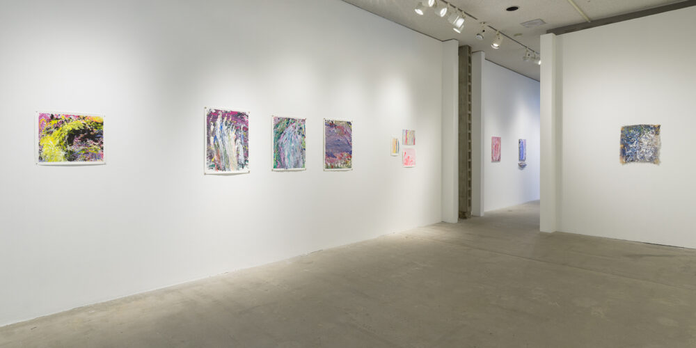 This is a color photograph of part of an exhibition of Vanessa McConnell's artworks at the Los Angeles Municipal Art Gallery. On the left wall are seven brightly colored abstract paintings on paper by Vanessa McConnell. On the far left is a painting featuring pink, yellow, and black paint in rough, dynamic brushstrokes. In the center of the wall are three paintings of equal size, consisting of various shades of blue, pink, purple, and green. On the right side of the wall are three small paintings grouped closely together in lighter shades of paint featuring mostly vertical brushstrokes. On the right wall is a single abstract painting on a piece of burlap featuring shades of blue, white, and green. Through an opening in this wall, two abstract paintings of roughly equal size can be seen on a wall in the distant background.